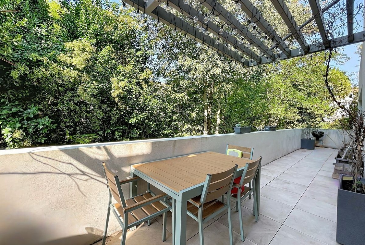 Terrasse carrelée avec pergola métallique anthracite garnie de guirlandes lumineuses, table de repas extérieure en bois composite avec quatre chaises assorties. Environnement arboré dense en arrière-plan, bac à plantes gris en bordure de terrasse.