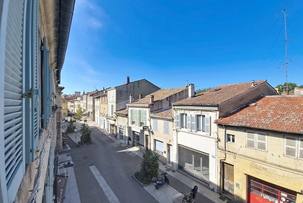 Vue depuis une fenêtre à volets bleus sur une rue semi-piétonne d'Avignon intra-muros, bordée de façades en pierre avec volets traditionnels et d'un commerce en rez-de-chaussée. En arrière-plan, les toits en tuiles romaines et un ciel bleu sans nuage caractérisent le cadre urbain historique du centre-ville d'Avignon.